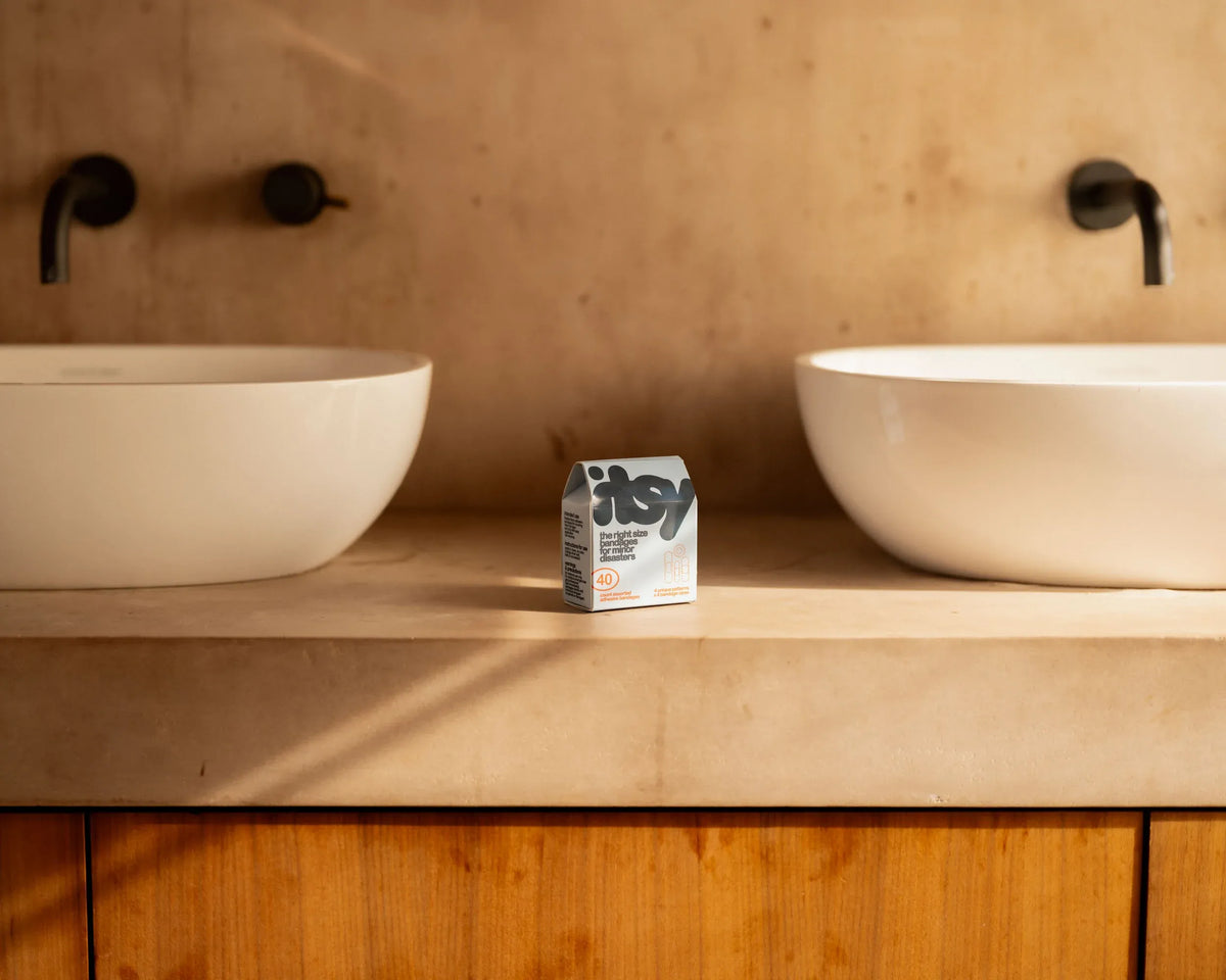 An itsy bandage box on a stone bathroom countertop between two vessel sinks